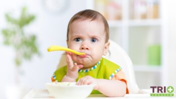 Bábätko sedí v detskej stoličke, drží lyžičku pri ústach a tvári sa váhavo. Má na sebe zelený podbradník, pred sebou má misku s jedlom, čo naznačuje, že práve skúša príkrmy.